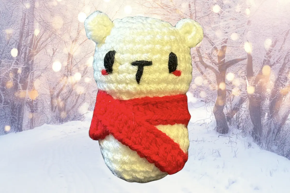 White crocheted polar bear cat toy wearing a red scarf against a snowy winter forest background.
